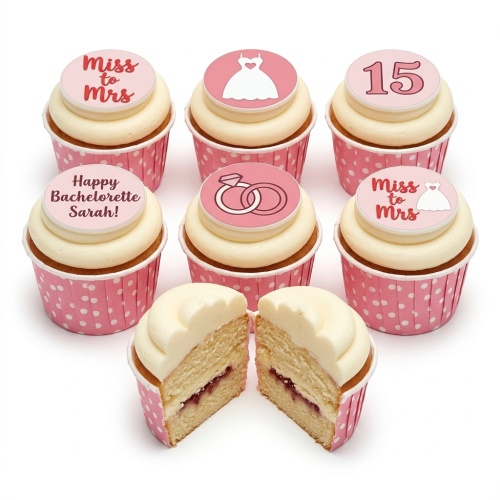 White cupcakes with pink polka dot wrappers and bachelorette themed toppers, including Miss to Mrs, wedding dress, rings, and custom age.