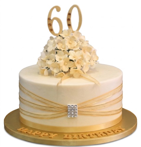 Elegant cream birthday cake with gold ribbons, a rhinestone buckle, a cluster of pale yellow sugar flowers, and a gold custom age topper.