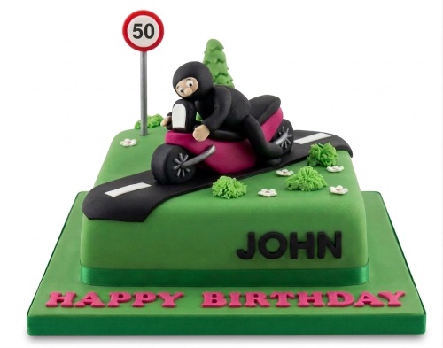 Green square birthday cake with a black biker on a pink motorbike, road, bushes, and a custom age speed limit sign.