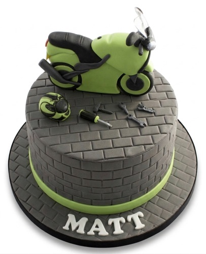 Round single-tier cake with a green motorcycle, helmet, and mechanic tools on a grey brick pattern, with a personalised name on the board.