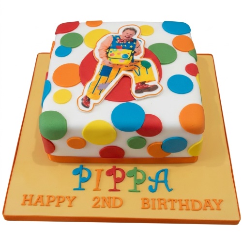 Square white cake with colourful polka dots and a Mr Tumble edible image on a yellow board.