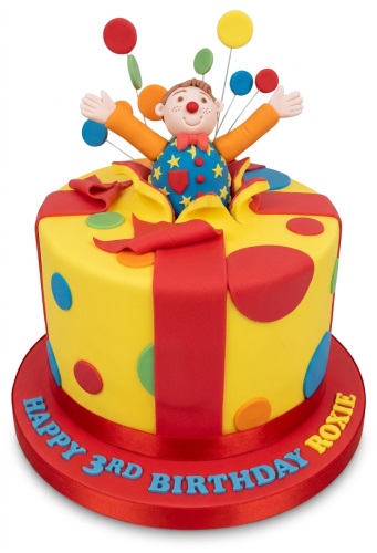 Yellow gift box cake with red ribbon, colourful polka dots, and a clown figure with balloons.
