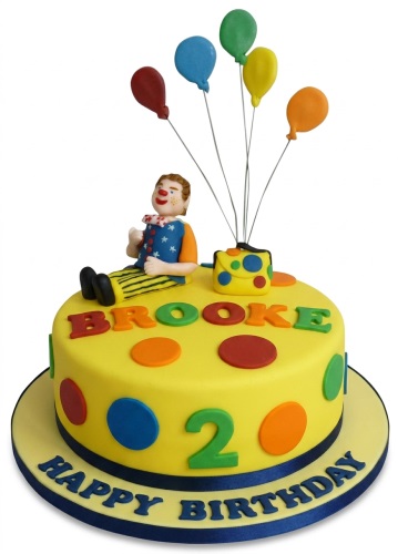 Yellow round birthday cake with a clown figure, colourful balloons, polka dots, and "Happy Birthday" message.