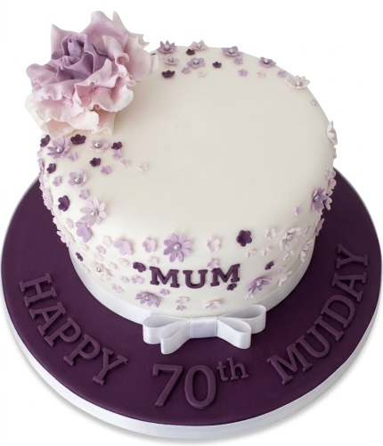 Round white cake with purple sugar flowers, a large ruffled flower, and a white ribbon bow.