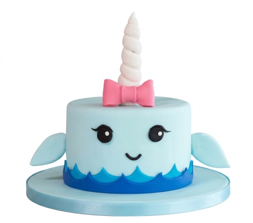 Light blue narwhal cake with a white glitter horn, pink bow, black eyes, and blue wave details.