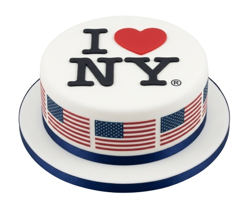 Round white cake with 'I Love NY' logo and red heart, surrounded by US flags and a blue ribbon.
