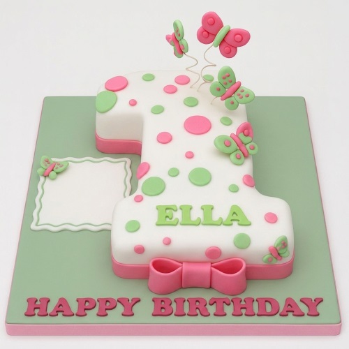 White number one cake with pink and green polka dots, butterflies, and a pink bow, for a girl's first birthday.