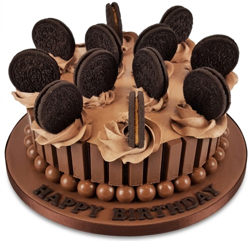 Round chocolate birthday cake decorated with Oreo cookies, Kit Kat bars, and chocolate malt balls, with 'Happy Birthday' on the board.