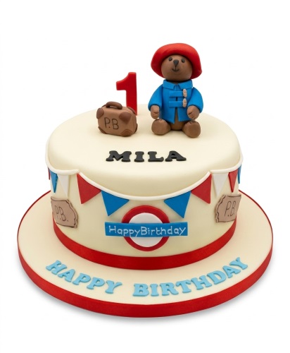 Round cream cake with Paddington Bear, suitcase, red number topper, and blue happy birthday text.