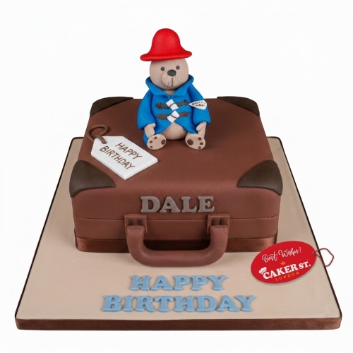 Paddington Bear figure on a brown suitcase birthday cake with a "Happy Birthday" tag and custom name.