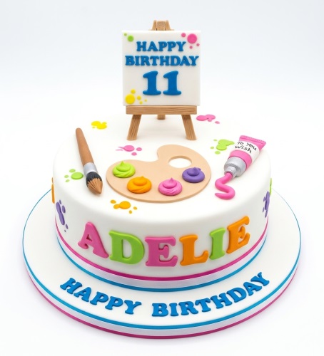 White round birthday cake with an art theme, featuring a paint palette, brush, easel with custom age, and personalised name.