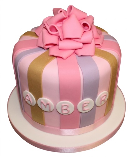 Round birthday cake decorated like a gift box with pink, lavender, and gold stripes, topped with a large pink fondant bow.