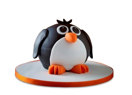 A round 3D penguin-shaped birthday cake with black, white, and orange icing, on a white board with an orange ribbon.