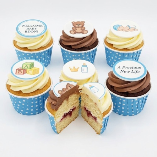Baby shower cupcakes for boys with blue polka dot cases, vanilla and chocolate frosting, and baby-themed edible toppers including a teddy bear, sleeping baby, ABC blocks, crown, and baby bottle.