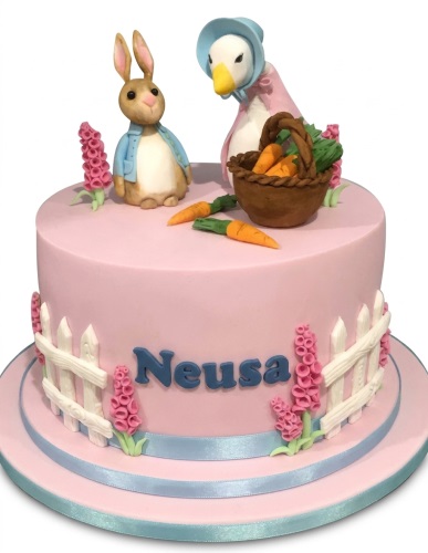 Pink round cake with Peter Rabbit and Jemima Puddle-Duck figures, carrots, white picket fence, and pink flowers.