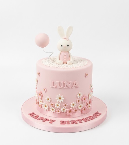 Pink round birthday cake with a white bunny topper, pink balloon, and white and pink flowers.