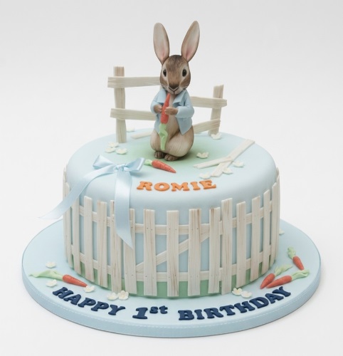 Round Peter Rabbit 1st birthday cake with fence, carrots, and blue ribbon.
