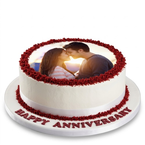 Round white anniversary cake with an edible photo of a couple on top, decorated with red velvet crumbs and a white ribbon, with 'Happy Anniversary' written in red on the board.