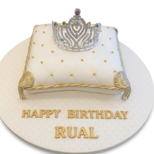 White pillow-shaped birthday cake with a silver tiara and gold details, featuring a custom message.