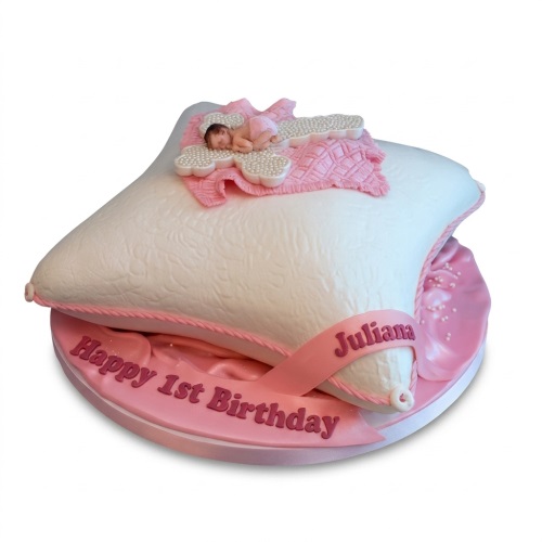 White pillow cake with sleeping baby figurine, pink blanket, pearls, and 'Happy 1st Birthday' ribbon.