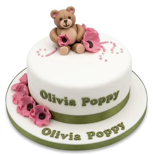 White fondant cake with a brown teddy bear topper, pink flowers, and green ribbon.