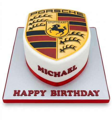 White shield-shaped cake with a large edible Porsche logo on top, red ribbon, and custom message on the board.