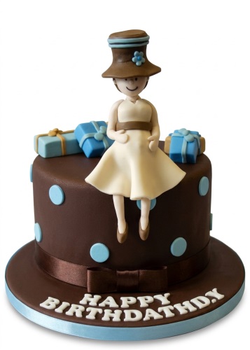 Round brown birthday cake with light blue polka dots, topped with a fondant lady figure in a cream dress and hat, surrounded by blue gift boxes, with 'Happy Birthday' written on the board.