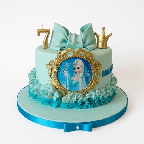 Light blue single-tier Frozen Elsa birthday cake with gold crown, bow, and custom age candle.