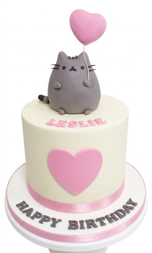 Round white birthday cake with a grey Pusheen cat topper holding a pink heart balloon and pink heart decoration.