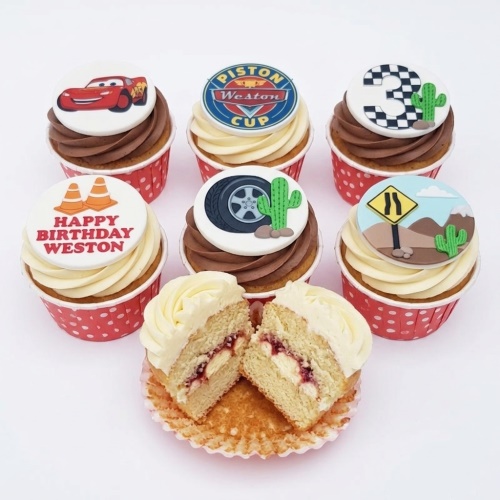 A selection of Cars theme birthday cupcakes with racing car, Piston Cup, road signs, tire, and cactus decorations, with one cupcake cut in half.