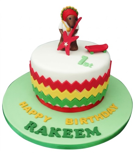 Round white birthday cake with reggae bear, guitar, skateboard, and Rasta zig-zag pattern.