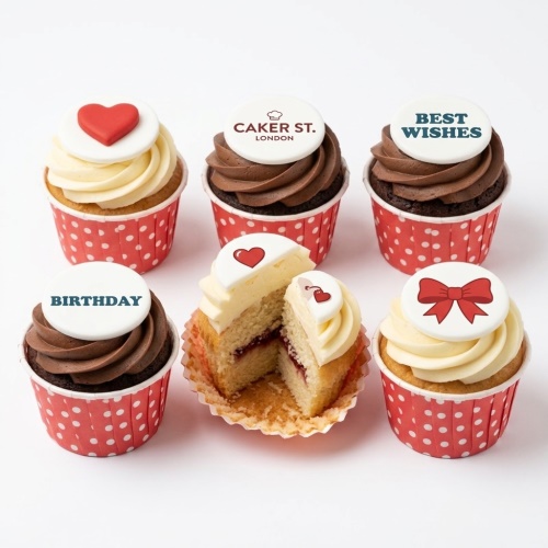 Six cupcakes with red polka dot cases, some with chocolate frosting, some with vanilla frosting. Toppers include a red heart, a red bow, and text 'BIRTHDAY', 'BEST WISHES', and 'CAKER ST. LONDON' logo. One cupcake is cut in half showing jam filling.