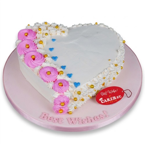 White heart-shaped cake with pink swirls, gold and blue sprinkles, and 'Best Wishes!' message.