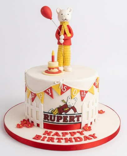 White round birthday cake with a Rupert Bear figure, red balloon, small cake with candle, Rupert Bear edible image, and 'Happy Birthday' message.