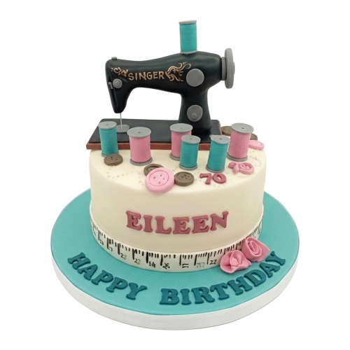 White birthday cake with black sewing machine, pink and teal spools, buttons, and measuring tape.