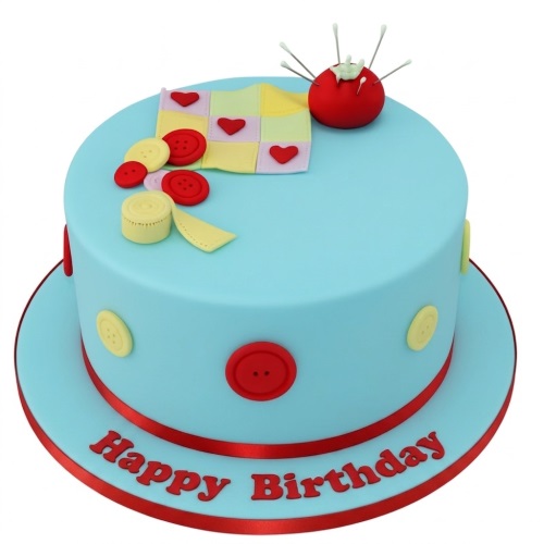 Light blue round cake with red and yellow buttons, a patchwork quilt, and a red pincushion with pins.