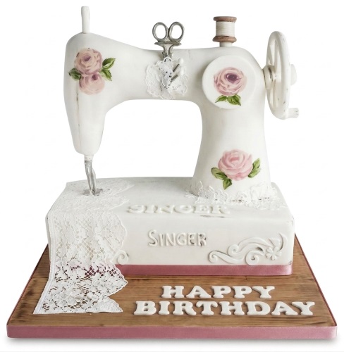 White vintage sewing machine birthday cake with pink roses and edible lace on a wooden base.