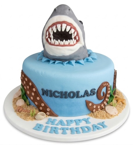 Single-tier blue fondant cake with a grey 3D shark head topper, octopus tentacles, and edible sand with seashells.
