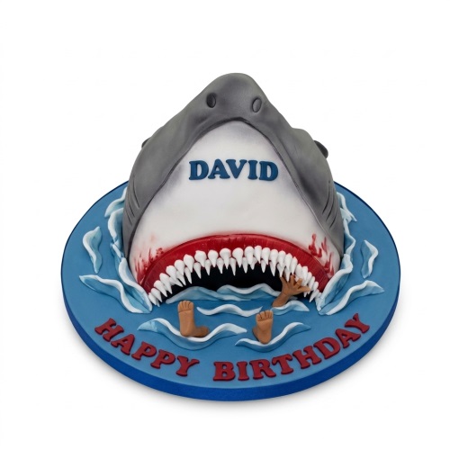 3D shark head birthday cake emerging from blue water with white teeth and red gums, personalised message.