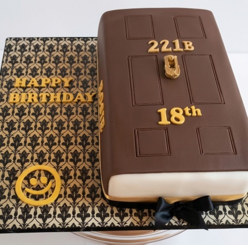 Brown fondant cake shaped like a door with '221B' and a door knocker. Cake board has a houndstooth pattern, 'Happy Birthday' text, and a smiley face. Includes a custom age.