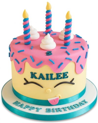 Round yellow kids birthday cake with pink drip, blue sprinkles, cartoon face, and spiral candles.