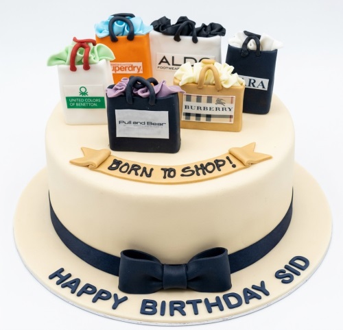 Round cream birthday cake with edible shopping bags, "Born to Shop!" banner, and dark blue bow.