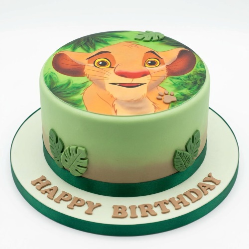 Round Simba The Lion King birthday cake with green and brown fondant, an edible Simba image, and green leaf decorations.