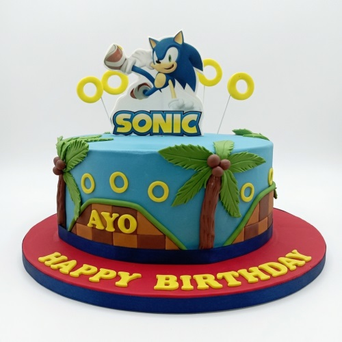 Round blue Sonic the Hedgehog birthday cake with character topper, golden rings, and palm trees.