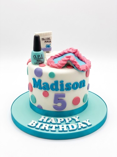 White round birthday cake with spa theme decorations, including nail polish, facial mask, and sleep mask, with colourful polka dots.