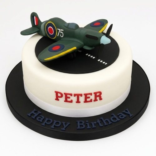 White round birthday cake with a green Spitfire aeroplane model on a black runway, with a custom name and message.
