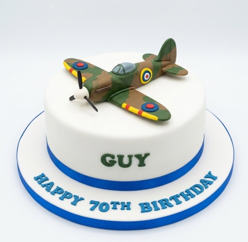 White birthday cake with a detailed camouflage Spitfire airplane model and blue ribbon.