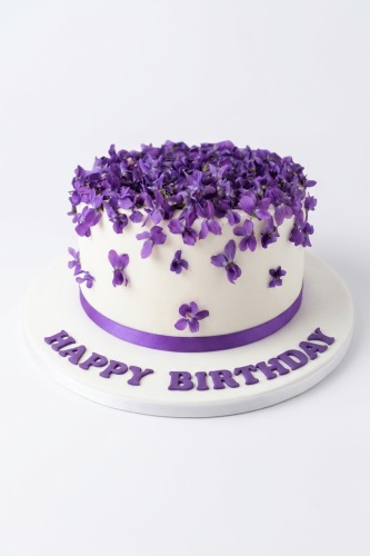 Round white birthday cake decorated with purple flowers and a purple ribbon, with 'Happy Birthday' written on the board.