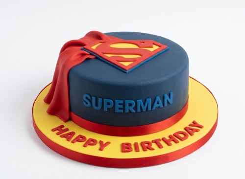 Round dark blue Superman birthday cake with red and yellow 'S' logo, red draped cape, and 'SUPERMAN' text. 'HAPPY BIRTHDAY' on yellow board.
