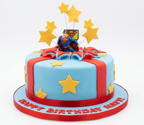 Round light blue Superman birthday cake with red ribbon, yellow stars, and Superman figurine topper.